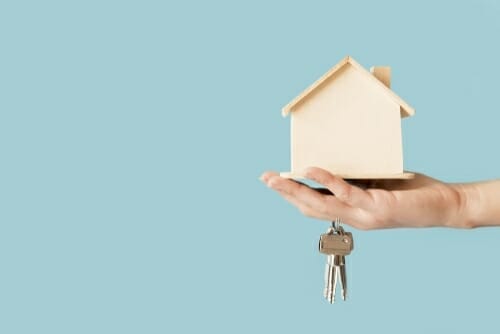 Close-up,Of,Hand,Holding,Keys,And,Wooden,House,Model,Against