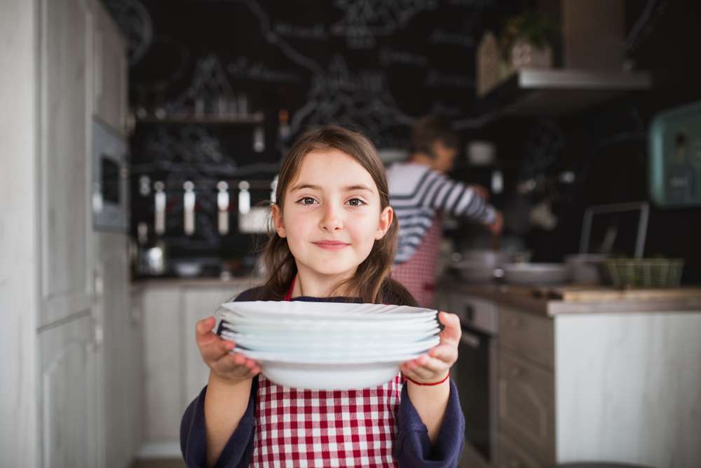 フランスのキッチンで、大人が後ろで作業をしている中、少女が積み重ねた皿を持ち、優しい笑顔でカメラに皿を差し出している。