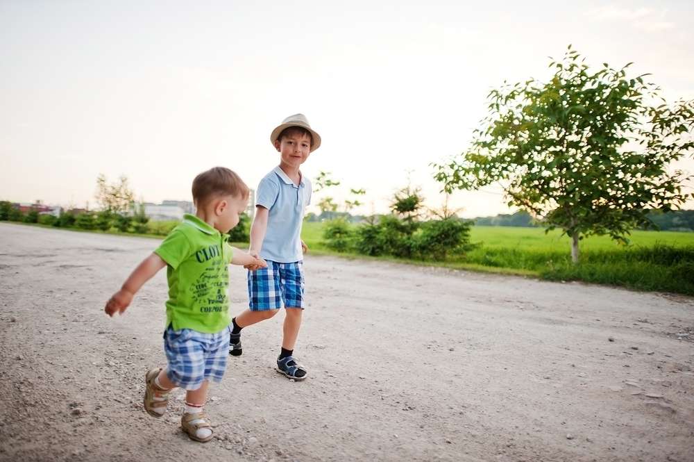 フランスの未舗装道路を歩いている 2 人の少年。1 人は水色のシャツ、青いチェック柄のショートパンツ、帽子を着用し、もう 1 人は緑のシャツと青いチェック柄のショートパンツを着用しています。道の両側には緑の野原と小さな木々が並んでいます。