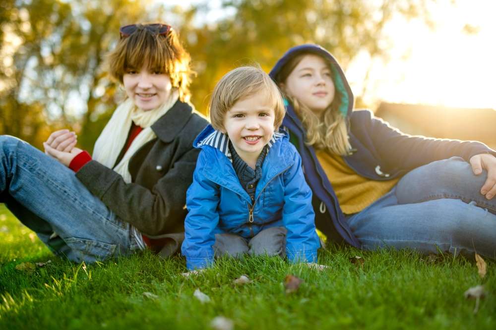 公園の芝生の上に座っている3人の子供。1人は灰色のコートを着ており、もう1人は真ん中で青いジャケットを着ており、3人目は紺色のコートを着てフードをかぶっています。背景には太陽の光が見え、フランスの午後を彷彿とさせます。