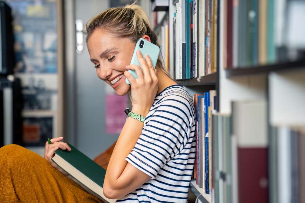 ある人が本棚に寄りかかって座り、フランスに関する本を開きながら、スマートフォンで話しながら微笑んでいます。