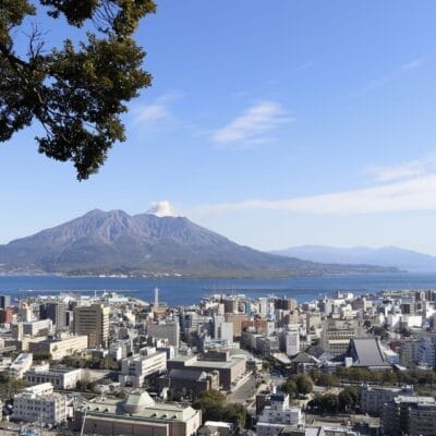 青空の下、大きな火山を背景にした活気に満ちた街並みは、フランス語教室マンツーマン鹿児島中央周辺の魅力を彷彿とさせます。左側の緑豊かな木々が自然の芸術性を高めています。.