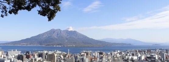 青空の下、大きな火山を背景にした活気に満ちた街並みは、フランス語教室マンツーマン鹿児島中央周辺の魅力を彷彿とさせます。左側の緑豊かな木々が自然の芸術性を高めています。.