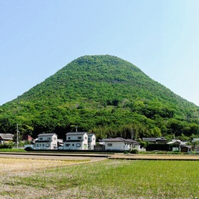 高松の澄み切った青空の下、住宅や畑の集落の背後に緑の円錐形の丘がそびえ立ち、香川の絵のような魅力を彷彿とさせます。.