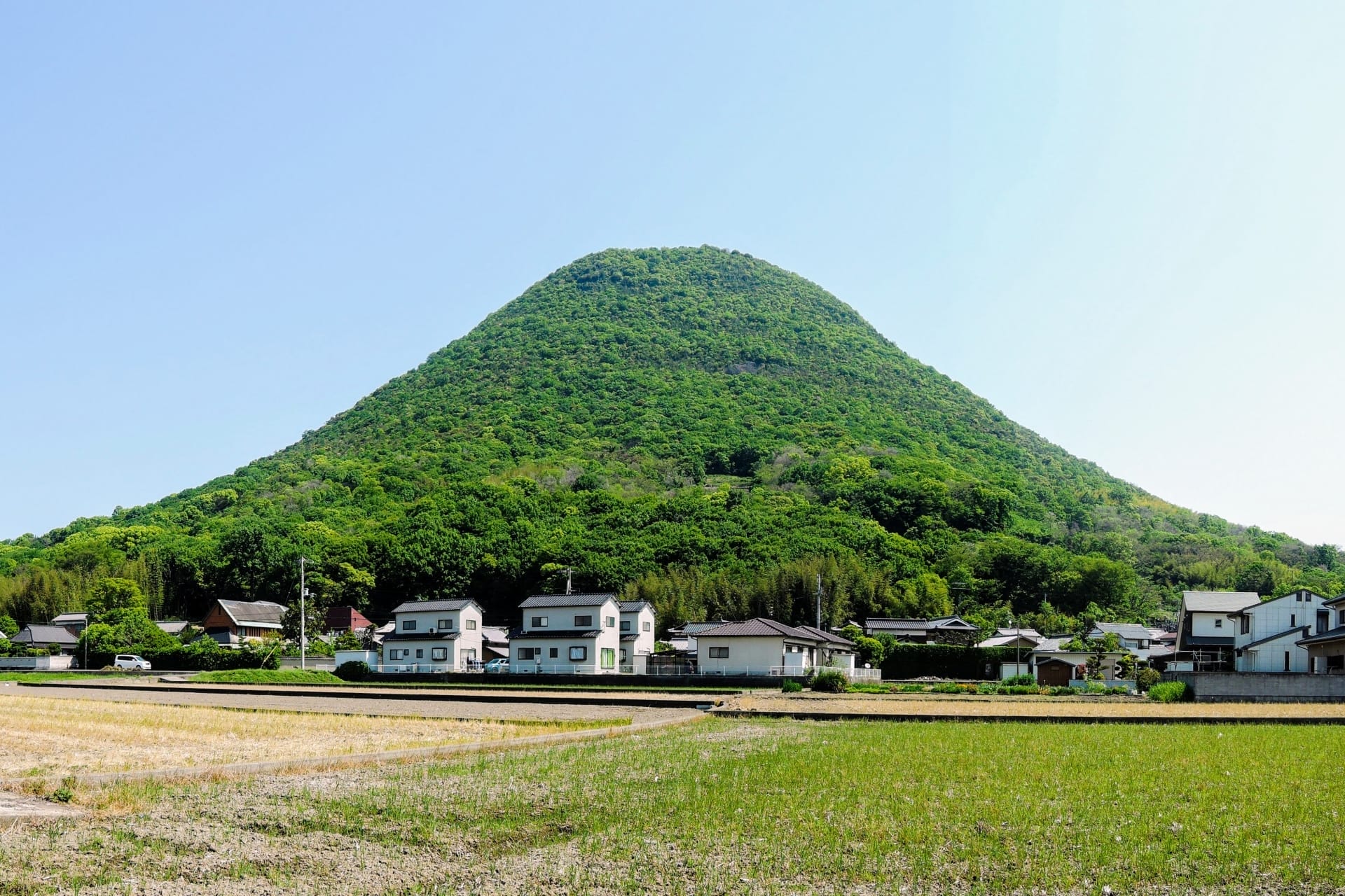 高松の澄み切った青空の下、住宅や畑の集落の背後に緑の円錐形の丘がそびえ立ち、香川の絵のような魅力を彷彿とさせます。.
