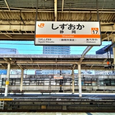 日本の静岡駅の空の電車のプラットホーム。頭上には日本語と英語の標識があります。静岡・三島が地域の魅力を添え、背景には優雅な建物がそびえ立っています。.