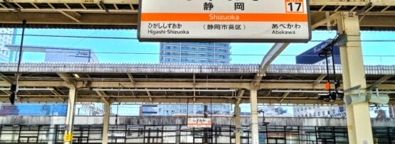 日本の静岡駅の空の電車のプラットホーム。頭上には日本語と英語の標識があります。静岡・三島が地域の魅力を添え、背景には優雅な建物がそびえ立っています。.