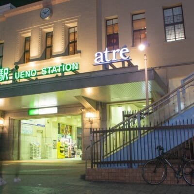 上野駅入口の夜景。明るい看板と柵のそばに駐輪された自転車。ぼやけた人々が通り過ぎ、動きを感じさせます。.