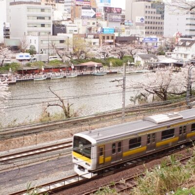 電車は飯田橋近くの都会の建物に囲まれ、桜が満開の川沿いの線路を走ります。そこではフランス語教室マンツーマンが文化的なタペストリーを加えています。.