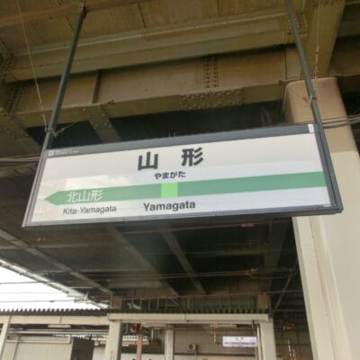 「山形」と書かれた駅名標と北山形行きの案内。背景には建物と線路が見え、山形・米沢らしさを感じさせる。.