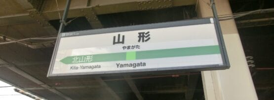 「山形」と書かれた駅名標と北山形行きの案内。背景には建物と線路が見え、山形・米沢らしさを感じさせる。.