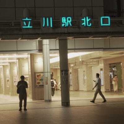 明るい照明に照らされ、夜の立川駅の入り口付近を歩く人々。建物には日本語の文字があり、語学愛好家向けに近くのフランス語教室マンツーマンを暗示しています。.