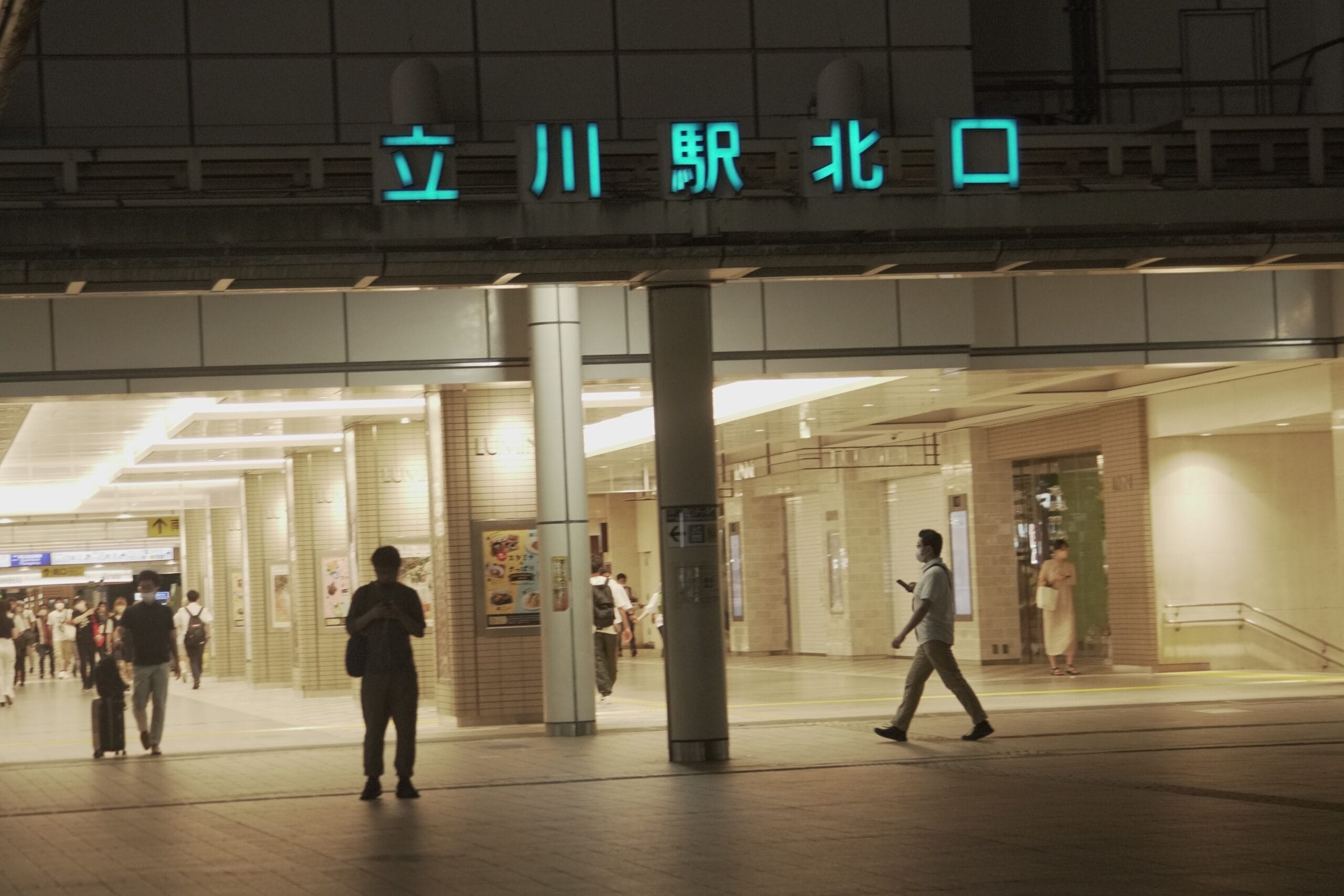 明るい照明に照らされ、夜の立川駅の入り口付近を歩く人々。建物には日本語の文字があり、語学愛好家向けに近くのフランス語教室マンツーマンを暗示しています。.