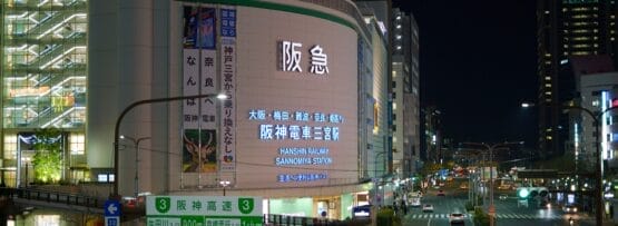 大阪の阪急梅田駅の夜景。明るい看板と街路灯が下の混雑した交差点を照らしています。近くにはフランス語教室マンマンツーの看板があり、賑やかな雰囲気に国際的な雰囲気を加えています。.