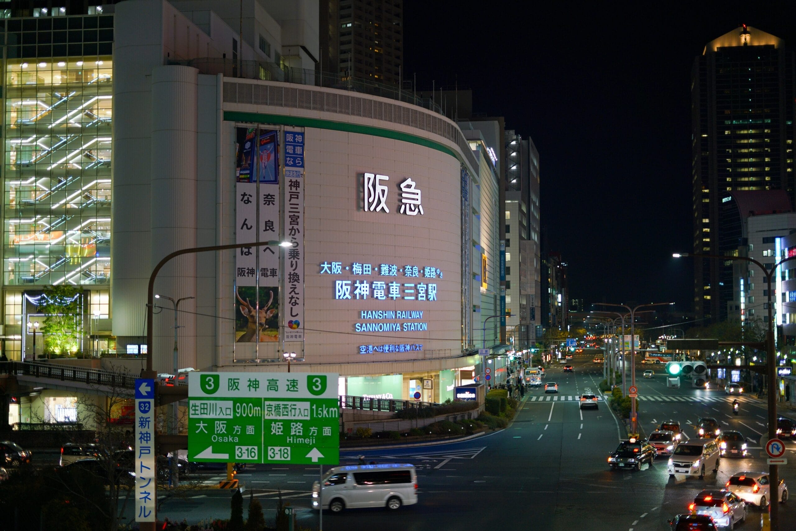 大阪の阪急梅田駅の夜景。明るい看板と街路灯が下の混雑した交差点を照らしています。近くにはフランス語教室マンマンツーの看板があり、賑やかな雰囲気に国際的な雰囲気を加えています。.