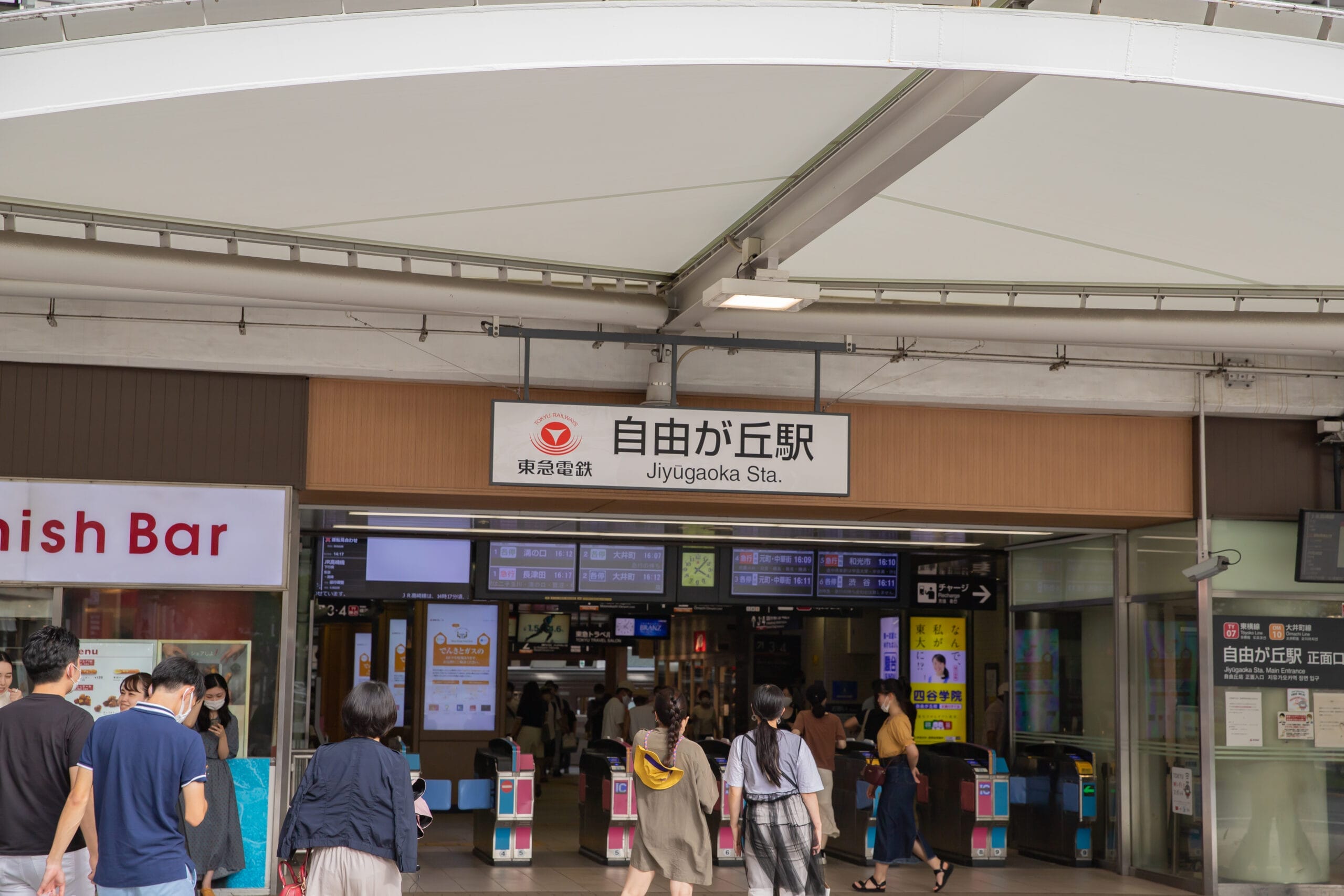 大きな白い天蓋の下、自由が丘駅の入り口に向かって歩く人々。入口の上には駅名標が見えており、次の目的地に向かう人や、近くにフランス語教室マンツーマンがある人を歓迎する雰囲気を醸し出しています。.