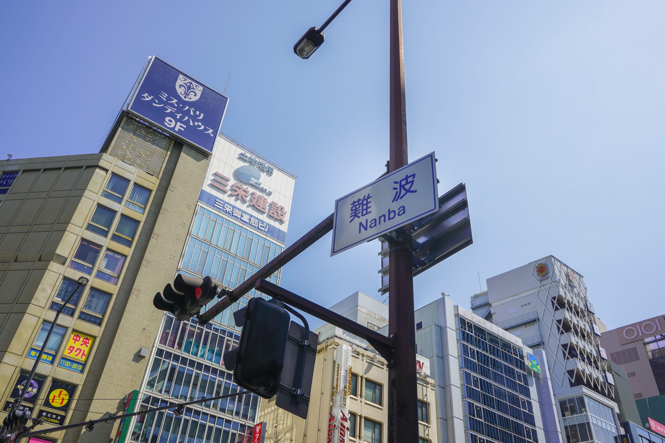 大阪の難波の街並み — 難波・なんば — 澄んだ青空の下、高層ビル、鮮やかな看板、信号機が立ち並ぶ。賑やかな街並みの中で、フランス語教室マンツーマンは、ダイナミックな都市のエネルギーの中で、個人に合わせた語学レッスンを提供します。.