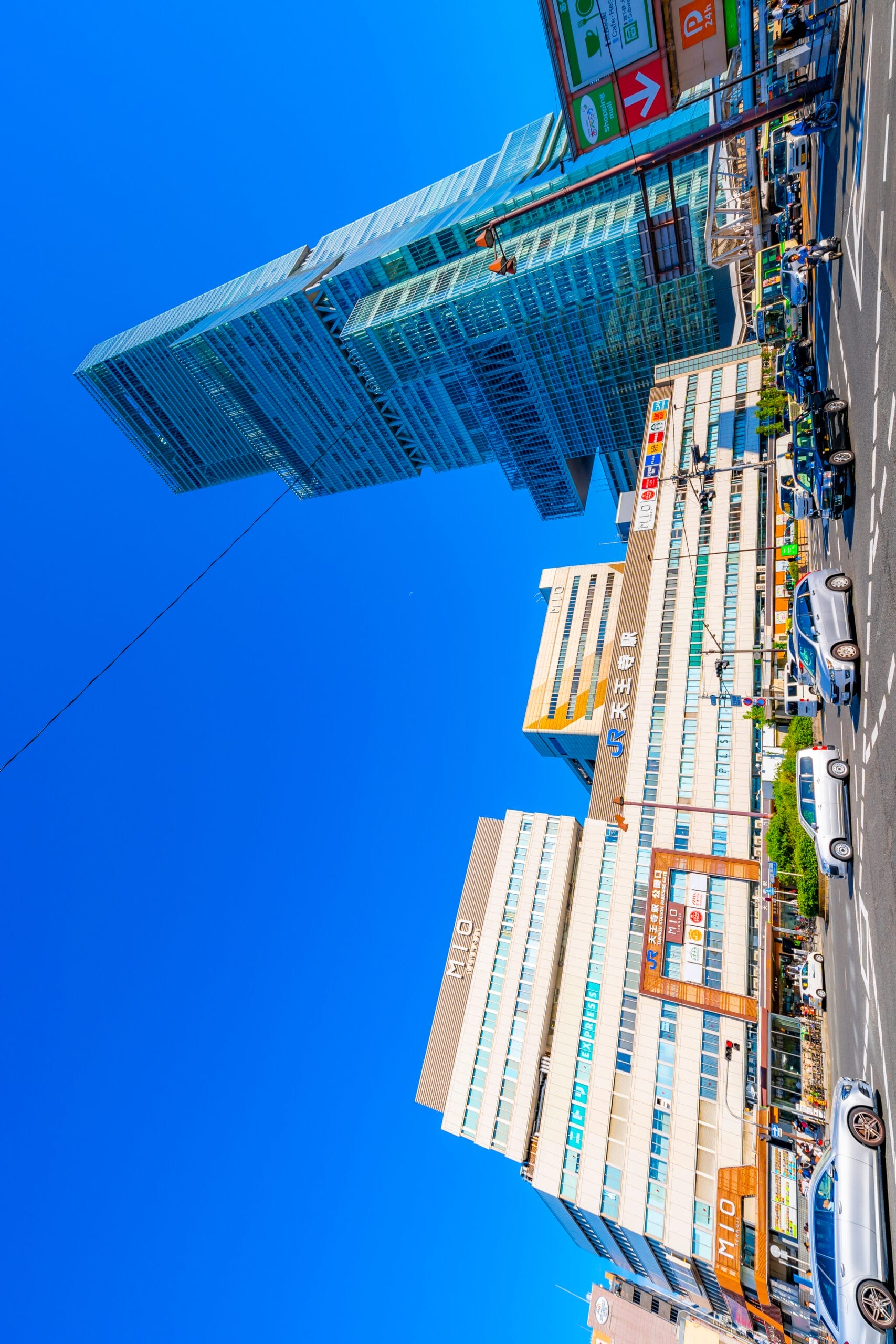 天王寺の澄んだ青空の下、高層ビルとにぎやかな街路風景。フランス語教室マンツーマンの広告を含む看板のあるさまざまな建物と、活気に満ちた喧騒の中を行き交うさまざまな車両が特徴です。.