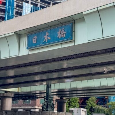 日本橋の近くには日本語の文字で書かれた標識のある高架道路が伸びており、背景には建設中の建物、その下には生い茂る木々が見える。.