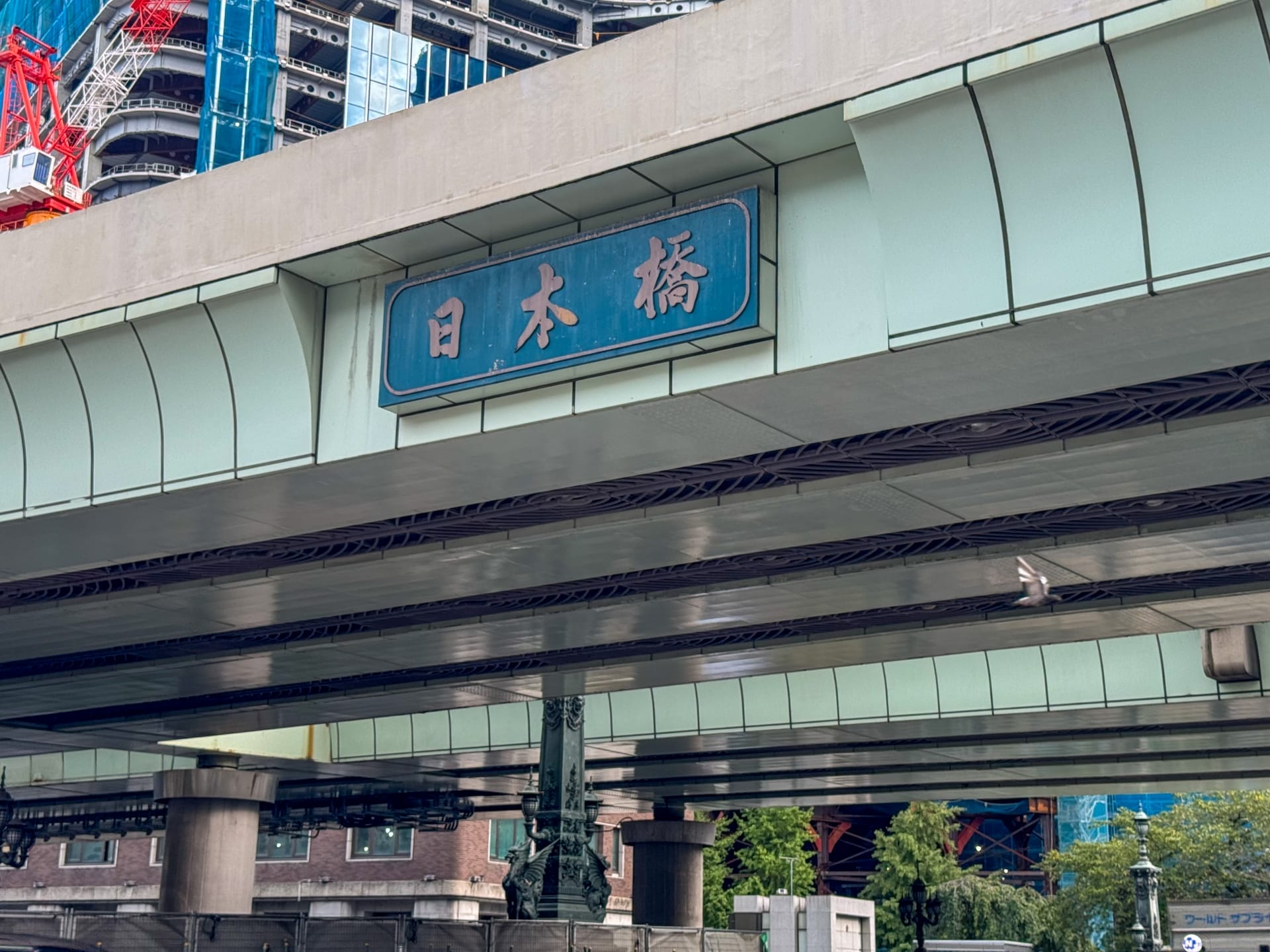 日本橋の近くには日本語の文字で書かれた標識のある高架道路が伸びており、背景には建設中の建物、その下には生い茂る木々が見える。. 日本橋の近くには日本語の文字で書かれた標識のある高架道路が伸びており、背景には建設中の建物、その下には生い茂る木々が見える。.