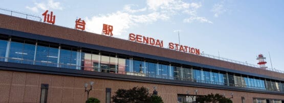 大きなレンガ造りの建物と、日本語と英語で書かれた目立つ標識が特徴的な仙台駅の外観。手前には澄んだ空の下の歩道と木々が広がり、宮城・仙台の活気あるエッセンスを捉えています。.