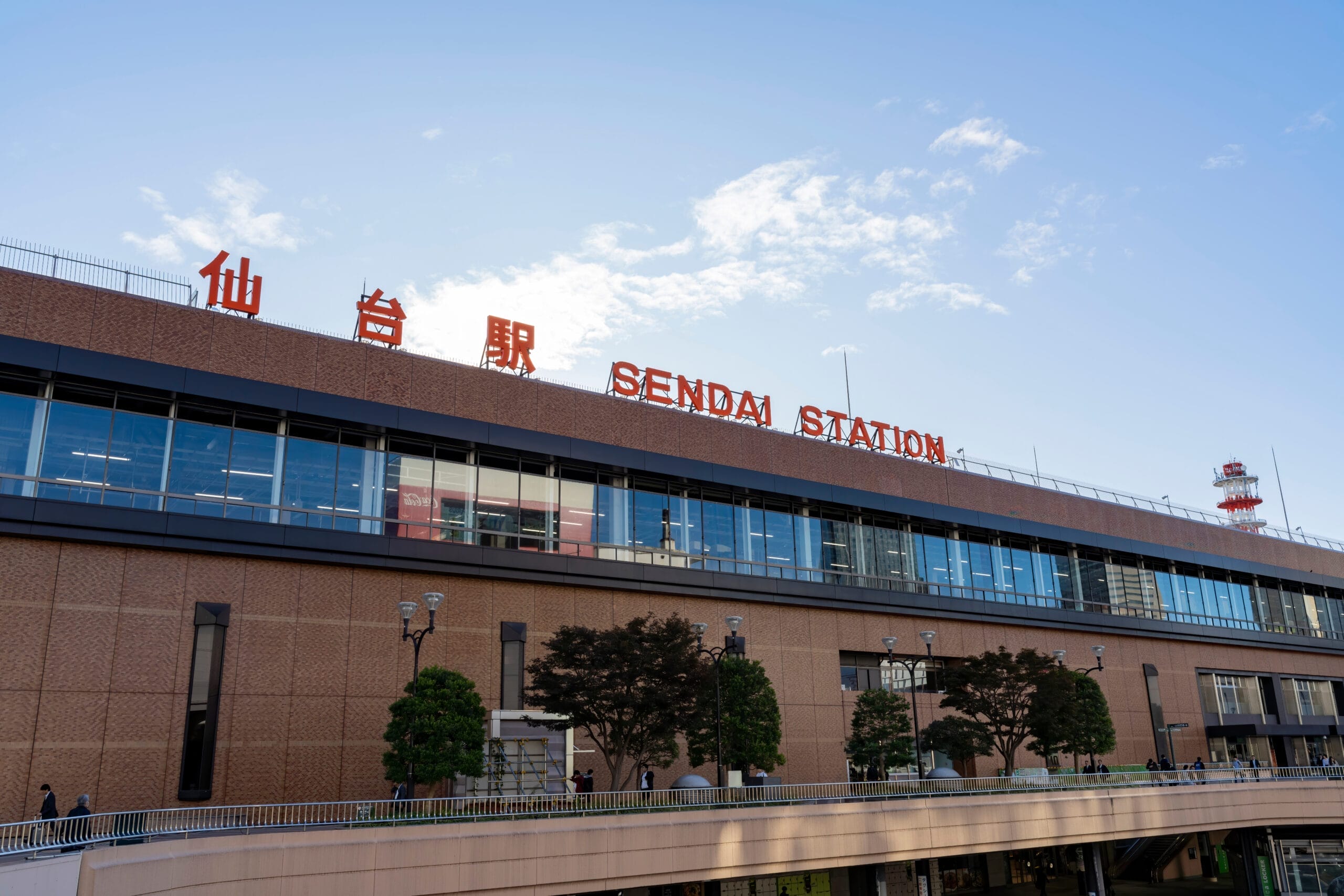 大きなレンガ造りの建物と、日本語と英語で書かれた目立つ標識が特徴的な仙台駅の外観。手前には澄んだ空の下の歩道と木々が広がり、宮城・仙台の活気あるエッセンスを捉えています。. 大きなレンガ造りの建物と、日本語と英語で書かれた目立つ標識が特徴的な仙台駅の外観。手前には澄んだ空の下の歩道と木々が広がり、宮城・仙台の活気あるエッセンスを捉えています。.