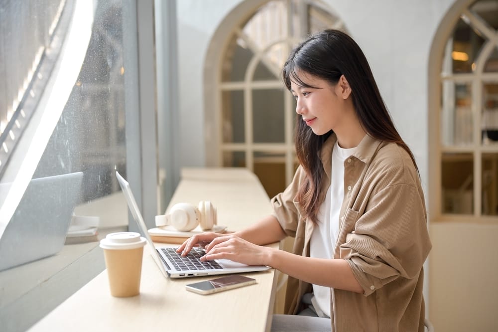 明るいカフェのテーブルで、コーヒーカップとスマートフォンを横に置いてノートパソコンを打つ女性。静かなフランスの朝のエッセンスを捉えています。.