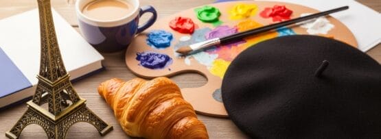 小さなエッフェル塔の模型、コーヒーカップ、クロワッサン、黒いベレー帽、ノート、画家のパレットと筆が木製のテーブルの上に並べられています。.