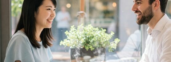 カフェのテーブルに向かい合って座り、コーヒーを飲みながら笑顔で会話する二人。テーブルの上にはノートとカラフルなカードが置かれ、背景には植物が置かれている。.